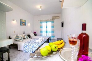 Quarto de hotel com uma cama, uma mesa com frutas e uma garrafa, e uma janela com cortinas.
