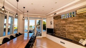 Un'ampia sala da pranzo con un lungo tavolo in legno e sedie, ampie finestre con vista sull'esterno e una TV integrata su un muro in pietra.