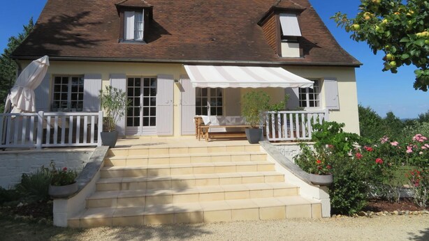 A two-story house with a covered porch, a staircase leading to the entrance, and a garden with flowers and a tree. - Le Coze (Saint-Martial-de-Nabirat)
