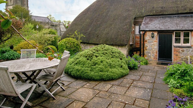 Une maison à toit de chaume avec un mur en pierre, un jardin agrémenté d’un arbuste rond et une table avec des chaises.