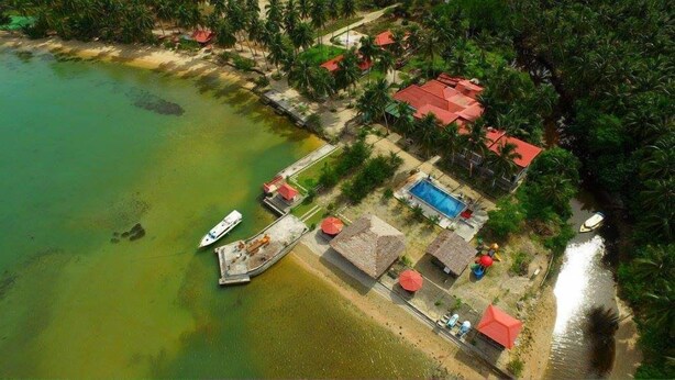 Vista aérea de una zona costera con piscina, un muelle con barcos y varios edificios de techo rojo rodeados de exuberante vegetación.