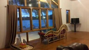 Amplia sala de estar con un ventanal que ofrece vista a una masa de agua. Hay una silla de madera y una mesita con un objeto decorativo.