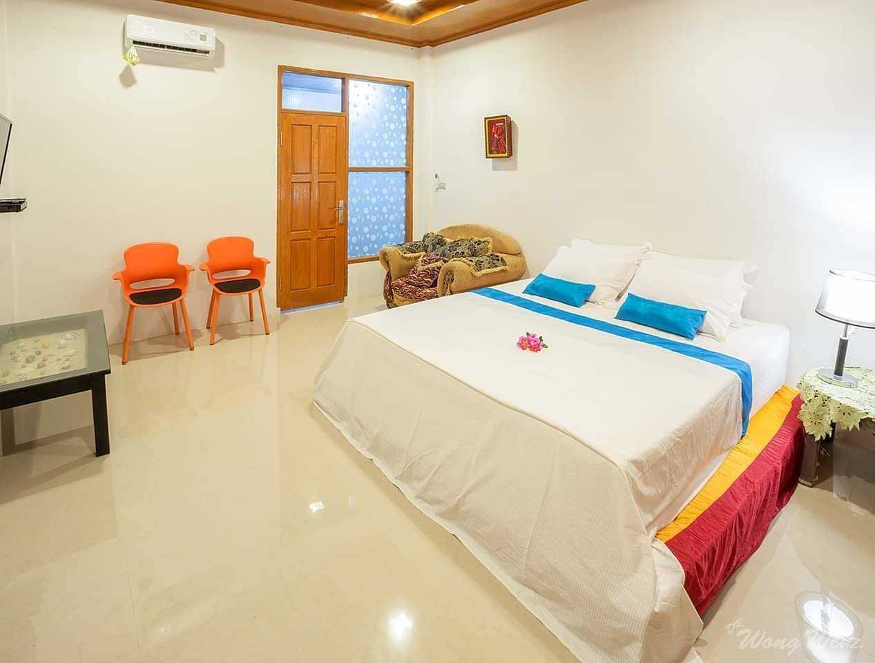 A bedroom with a large bed, two orange chairs, a wooden door, and a small table with a glass top.
