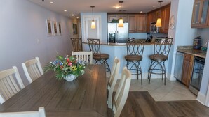 A dining area with a wooden table and chairs, a flower centerpiece, and a kitchen in the background. - MVG 1510 Oceanfront, Penthouse (North Myrtle Beach)