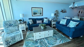 A living room with a blue sofa set, a patterned armchair, a coffee table, and a painting on the wall. - MVG 1510 Oceanfront, Penthouse (North Myrtle Beach)