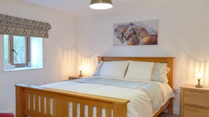 A bedroom with a wooden bed, bedside lamps, a window with curtains, a painting on the wall, and a red chair. - Hen Llaeth - UK11209 (Cwmsychpant, near Lampeter)