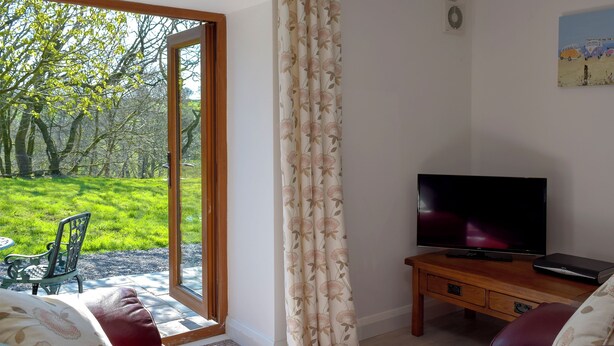 A living room with a television on a wooden stand, a sofa, and a large window with curtains. - Hen Llaeth - UK11209 (Cwmsychpant, near Lampeter)