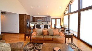 A spacious living room with a large window, a sofa, a coffee table, and a kitchen in the background. - The Summit - 3 bedroom 2.5 bathroom home - walk to town (Estes Park)