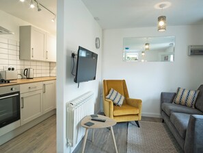 Eine moderne Küche mit weißen Schränken, einem eingebauten Ofen und einer Mikrowelle. Ein gelber Sessel mit einem gemusterten Kissen, ein graues Sofa und ein kleiner runder Tisch mit einer Fernbedienung sind ebenfalls vorhanden.
