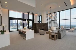 Relax in a spacious living area with a central seating area, kitchen, and floor-to-ceiling windows offering city views.