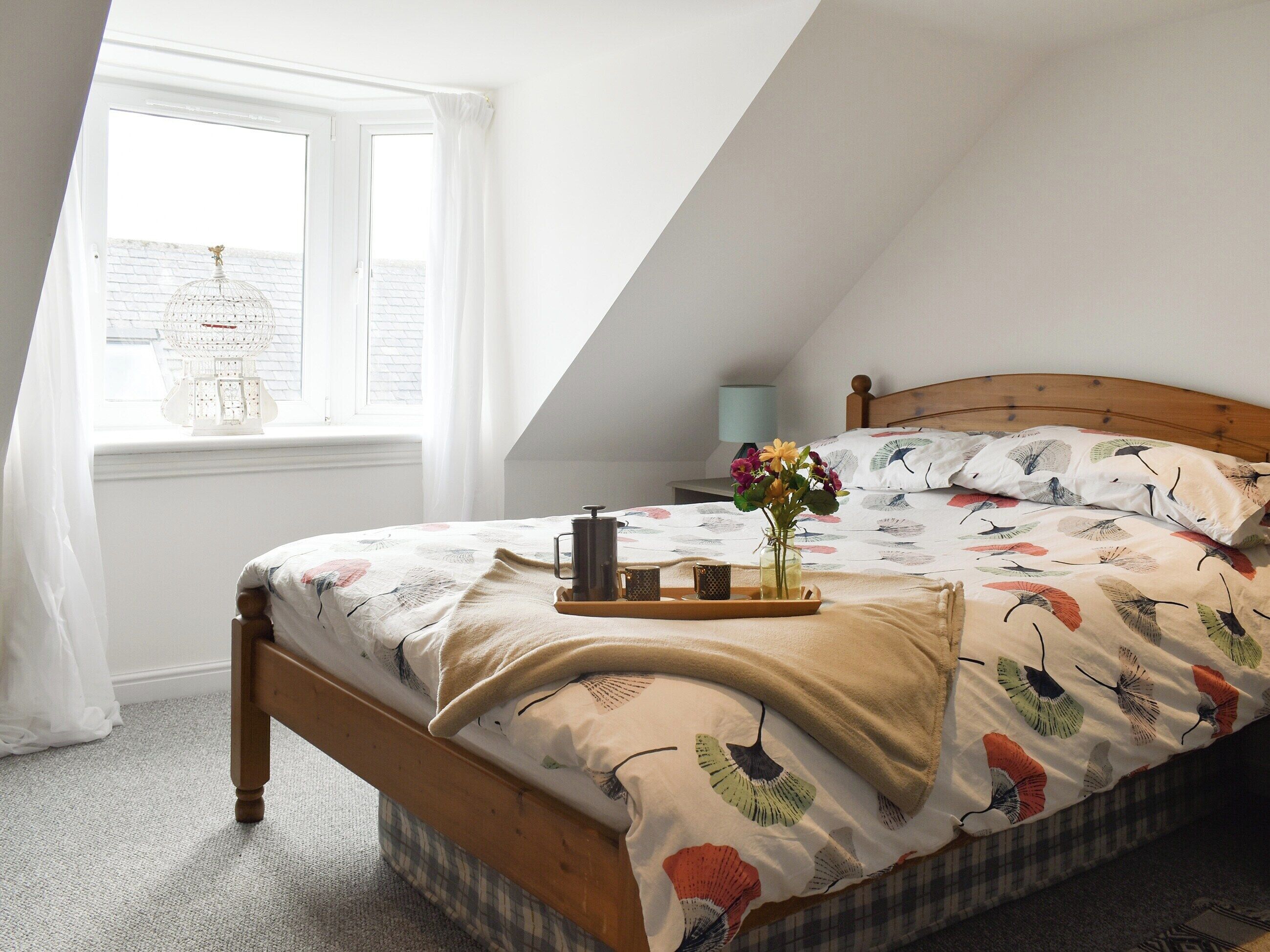 A bedroom with a sloped ceiling, a wooden bed frame, a window with a white curtain, a bedside table with a lamp, and a tray with a vase of flowers on the bed.