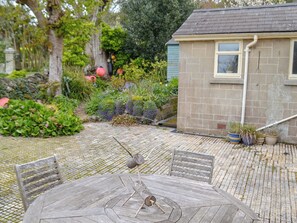 Un espace extérieur rustique aménagé avec une table et des chaises en bois rond, entouré d’un jardin où poussent différentes plantes, et avec un petit bâtiment visible au fond.