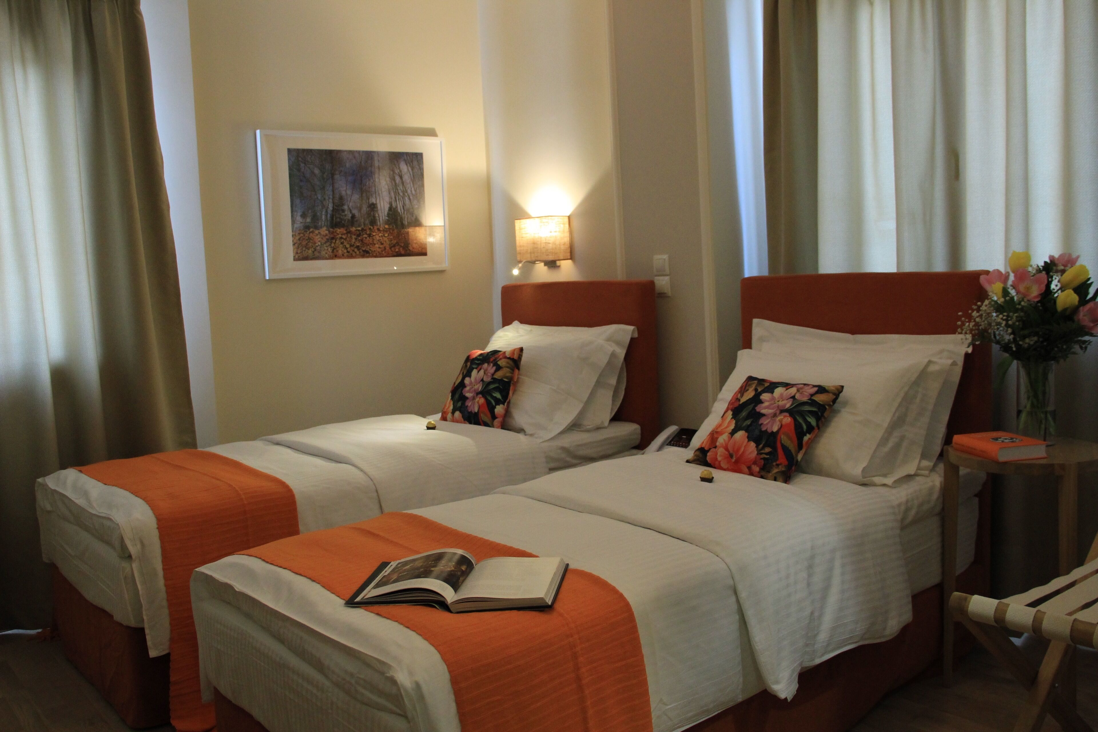 Una camera d'albergo con due letti singoli, un comodino con una lampada, un quadro appeso al muro e un tavolino con un vaso di fiori.