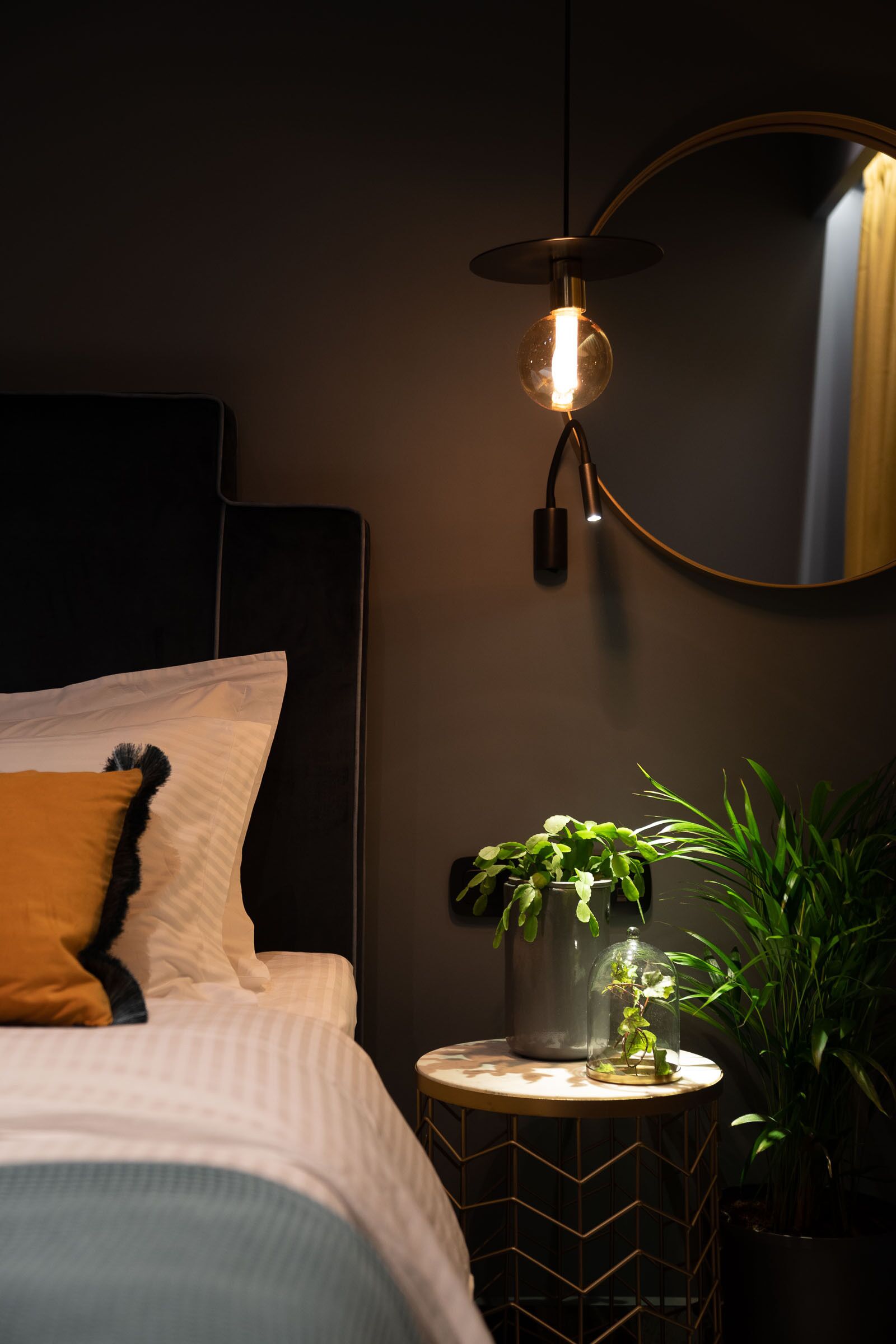 Ein ordentlich bezogenes Bett mit einem weißen und orangefarbenen Kopfkissen, ein kleiner runder Tisch mit einer Pflanze und eine Wandlampe.