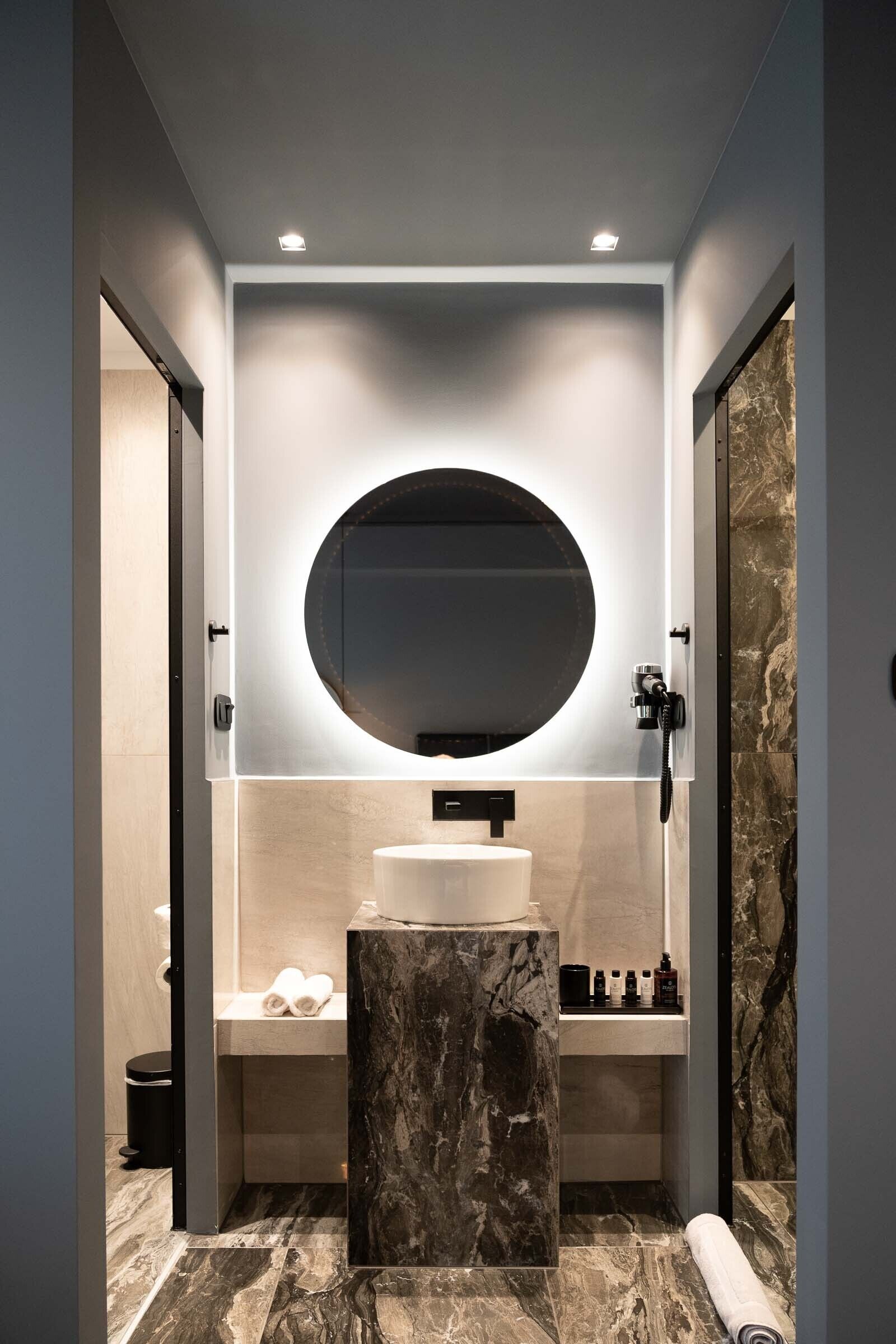 A modern bathroom with a round mirror, a marble pedestal sink, and a handheld showerhead.