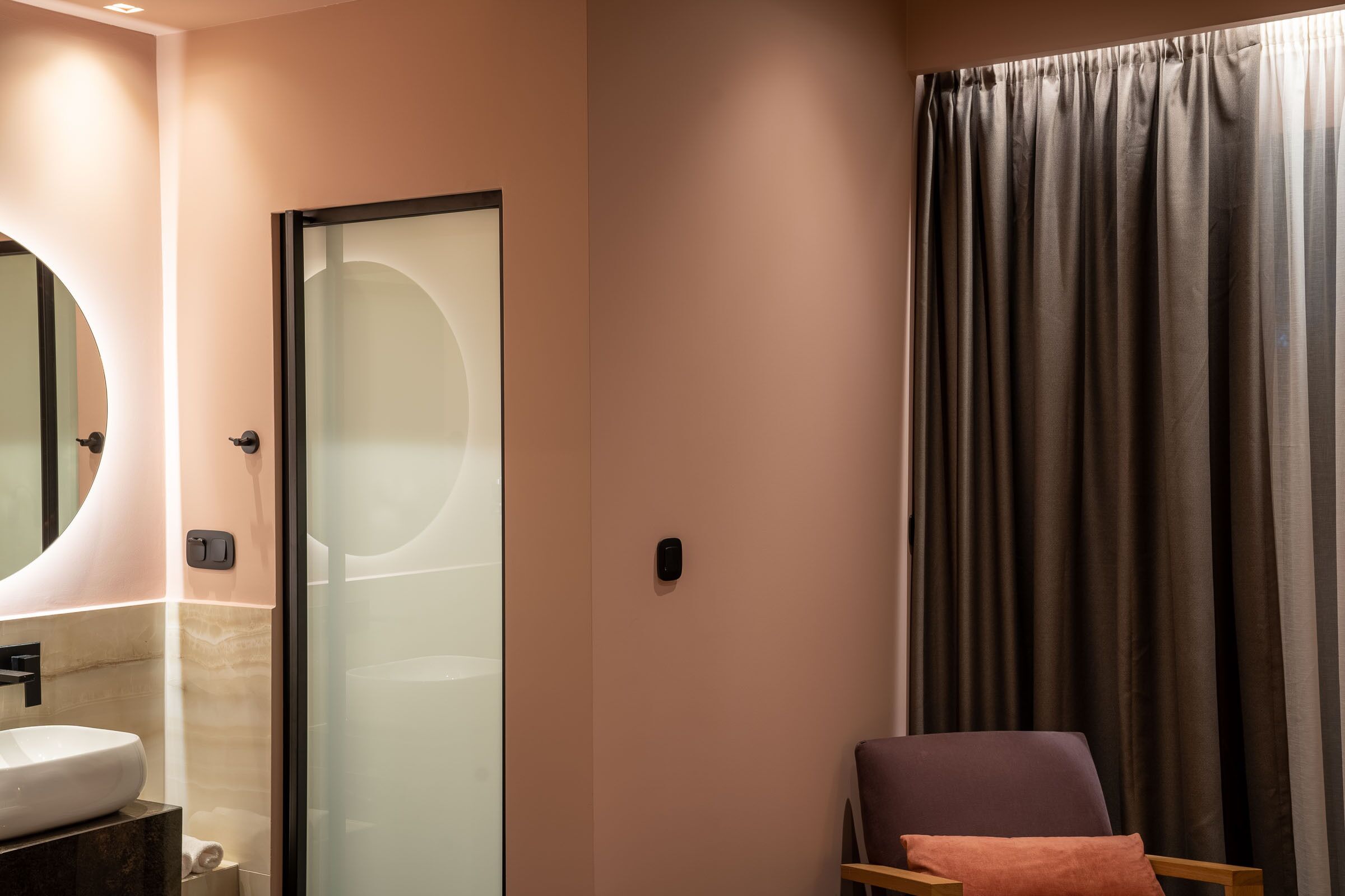 A modern bathroom with a glass shower enclosure, a round mirror, and a wooden chair with a cushion.
