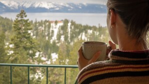 Une personne tenant une tasse, dominant un paysage montagneux avec des arbres et une étendue d’eau.