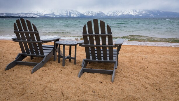 Deux fauteuils Adirondack sur une plage de sable, avec vue sur un lac et des montagnes enneigées en arrière-plan.
