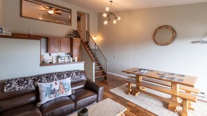 Un salon chaleureux avec un canapé en cuir marron, une table basse en bois, une table à manger en bois avec des bancs, un ventilateur de plafond, un miroir mural et un escalier menant à une autre pièce.