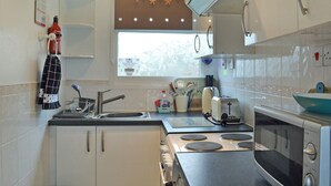 Una cocina compacta con estufa, microondas y fregadero. Hay una ventana con cortina y un estante pequeño sobre el fregadero.