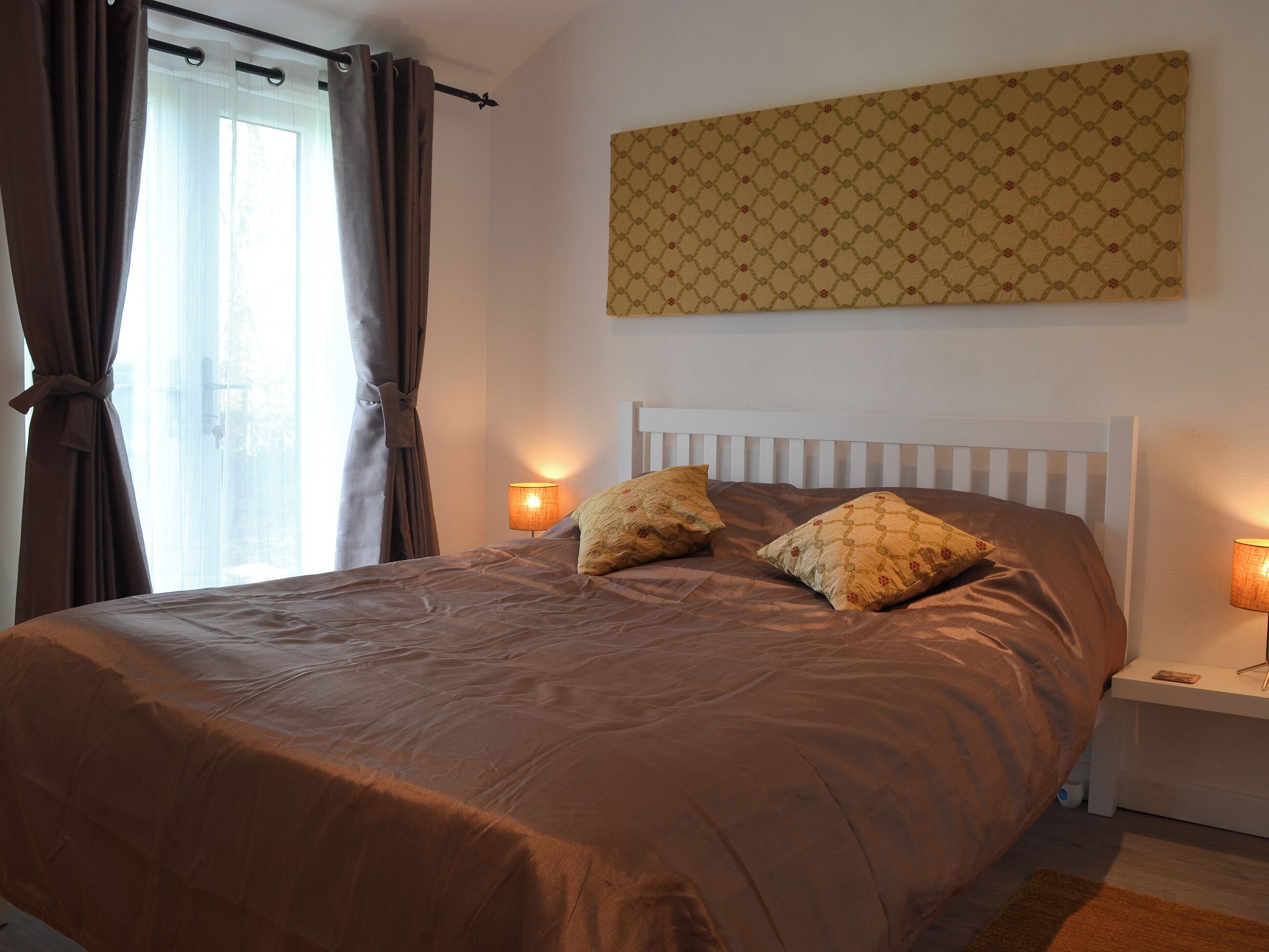 Un lit bien fait, avec une couverture marron et deux coussins décoratifs, une tête de lit blanche et une lampe fixée au mur de chaque côté.