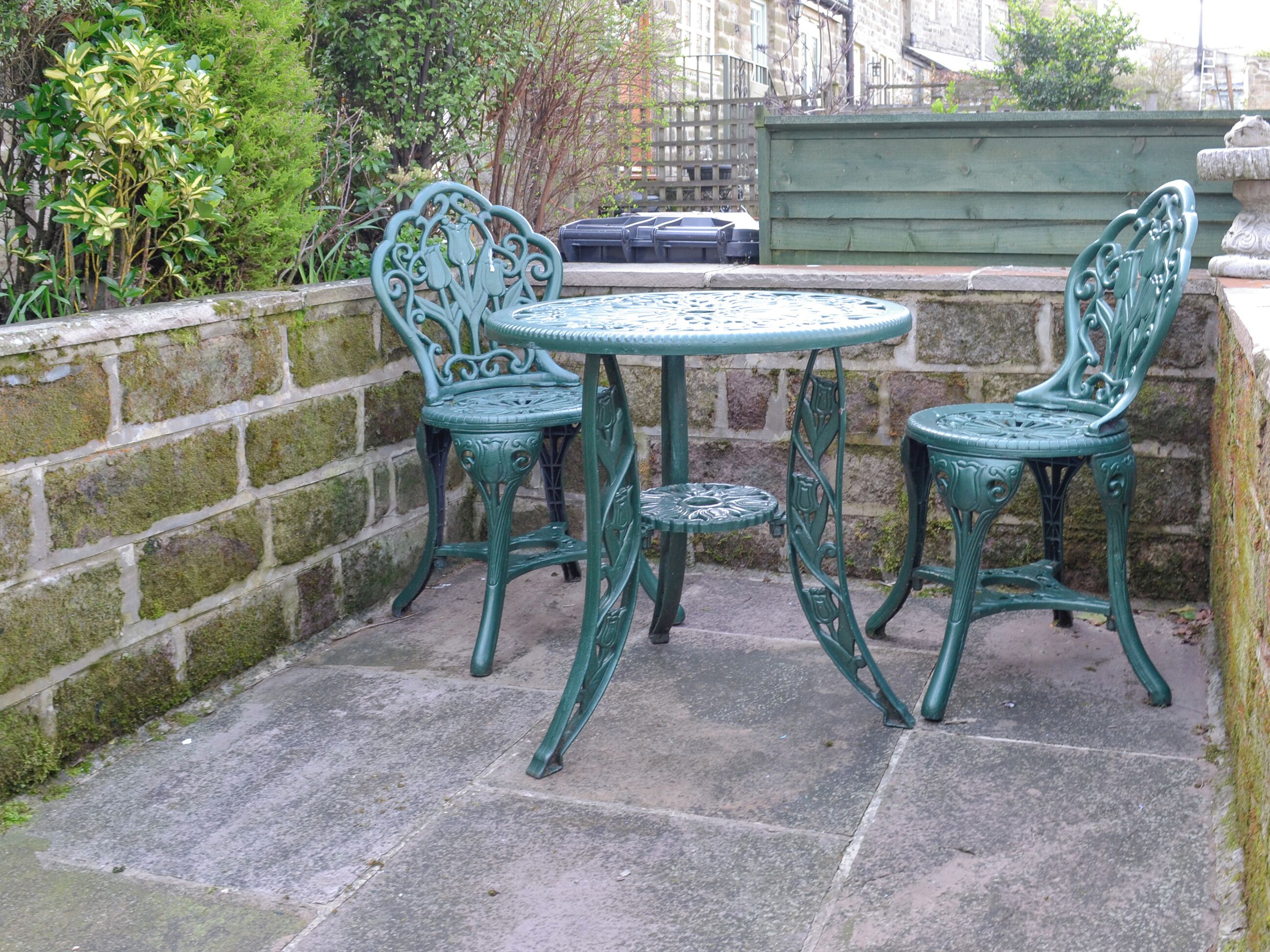 A patio with a round table and two chairs, surrounded by a stone wall and greenery.