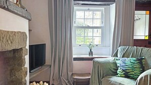 A cozy room with a stone fireplace, a television, a window with curtains, a bench, and a green sofa with a fern-patterned pillow. - 2 bedroom accommodation in Bewerley, near Pateley Bridge (Bewerley, near Pateley Bridge)