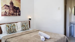 A hotel room with a bed, a painting of a cathedral, a bathroom with a toilet and sink, and a door leading to another room.