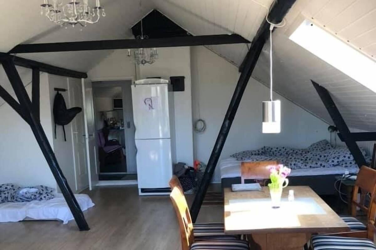 A cozy attic bedroom with a sloped ceiling, a wooden dining table with chairs, a bed, and a kitchenette area.
