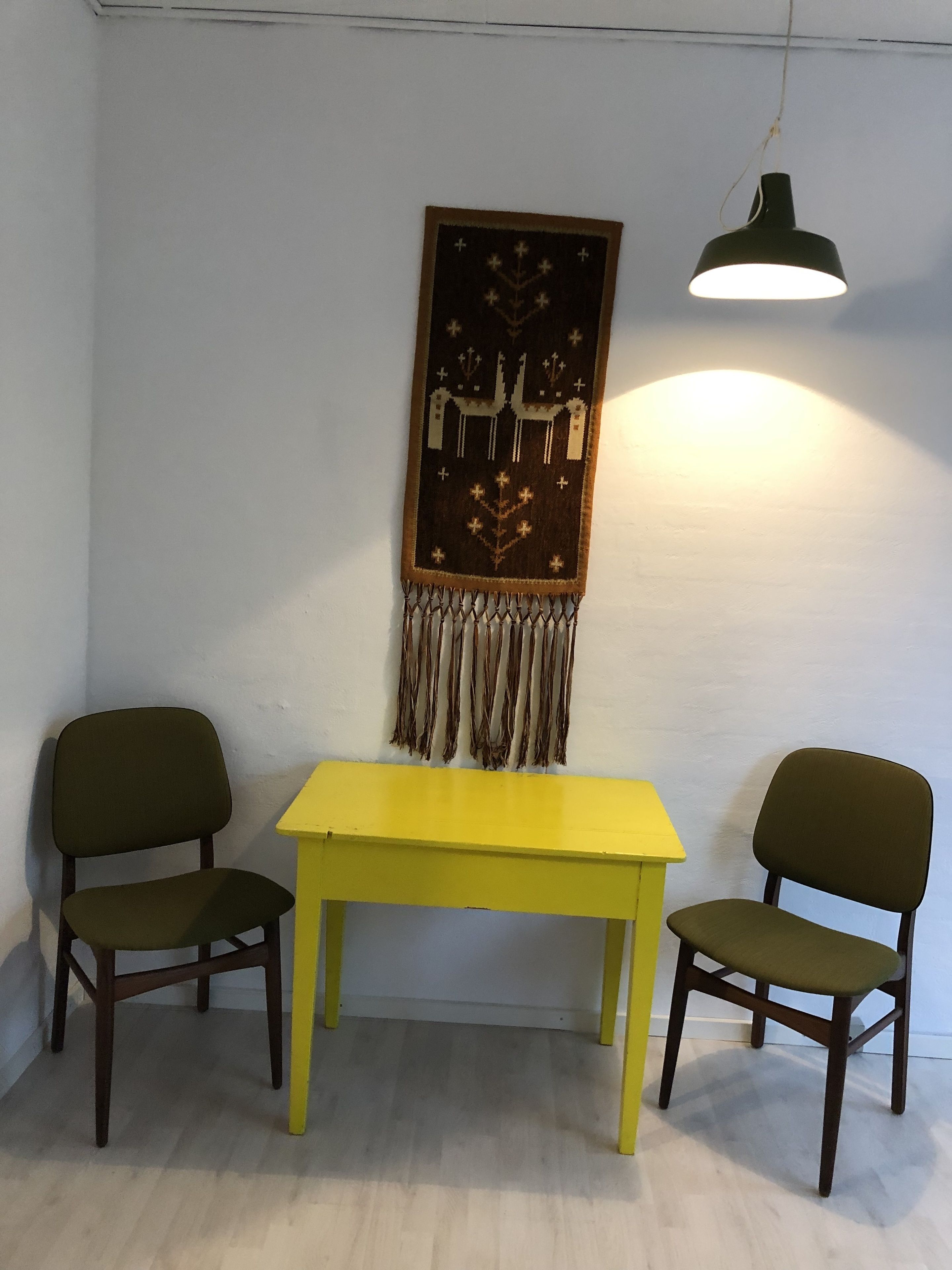 Una stanza con un tavolo giallo e due sedie, un arazzo a parete e una lampada a sospensione.