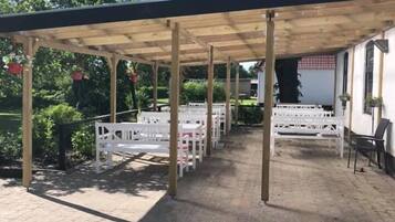 Un espacio al aire libre techado, con piso de ladrillo, vigas de madera y bancos blancos.