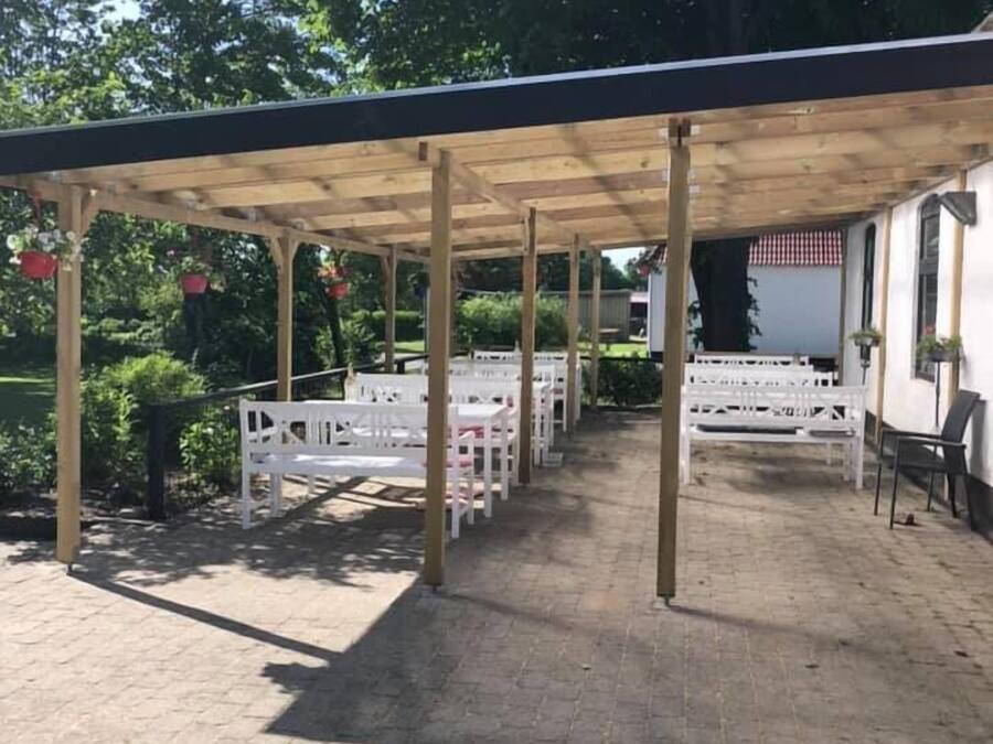 Un espacio al aire libre techado, con piso de ladrillo, vigas de madera y bancos blancos.