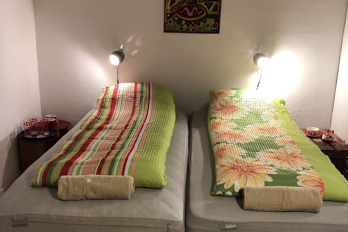 Two single beds with striped bedding, a wall-mounted painting, bedside lamps, and a small table with a cup and saucer.