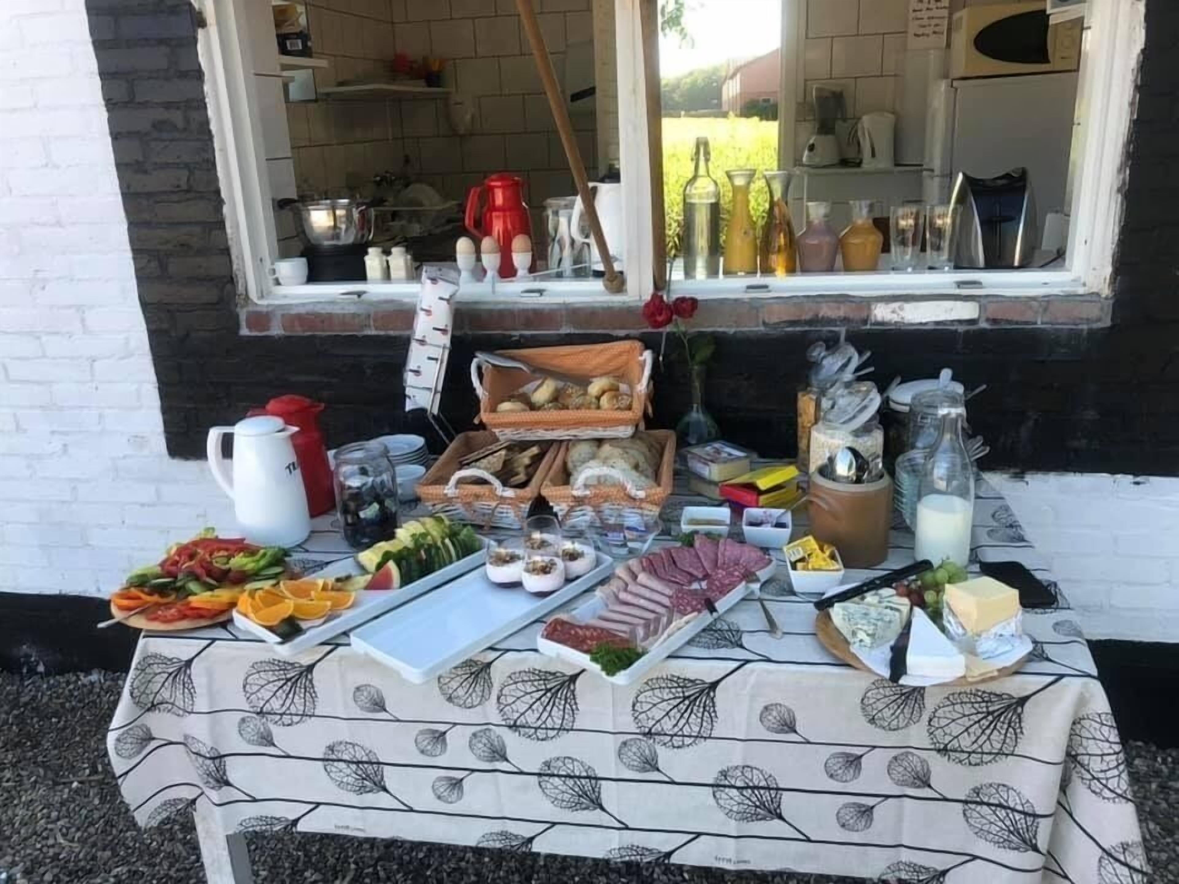 A table set outdoors with various food items and condiments, a coffee pot, and a window view of a kitchen.