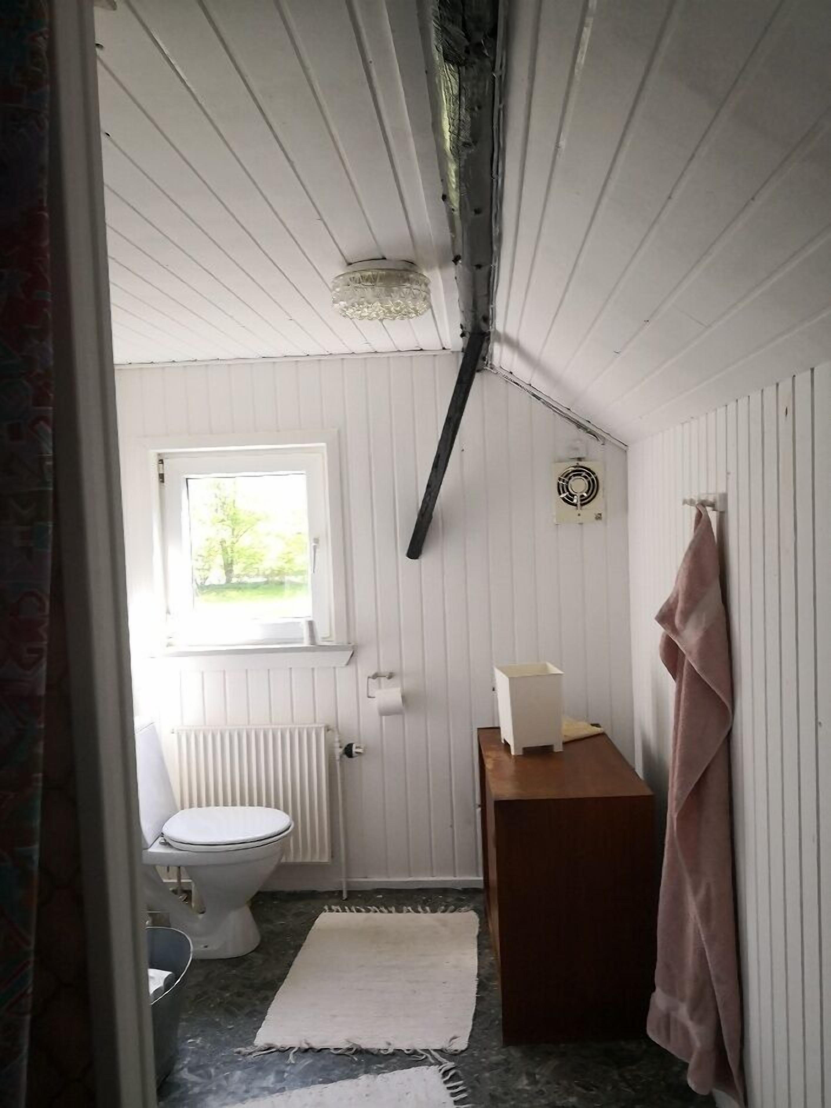 A small, compact bathroom with a toilet, a radiator, a window, and a wooden cabinet.