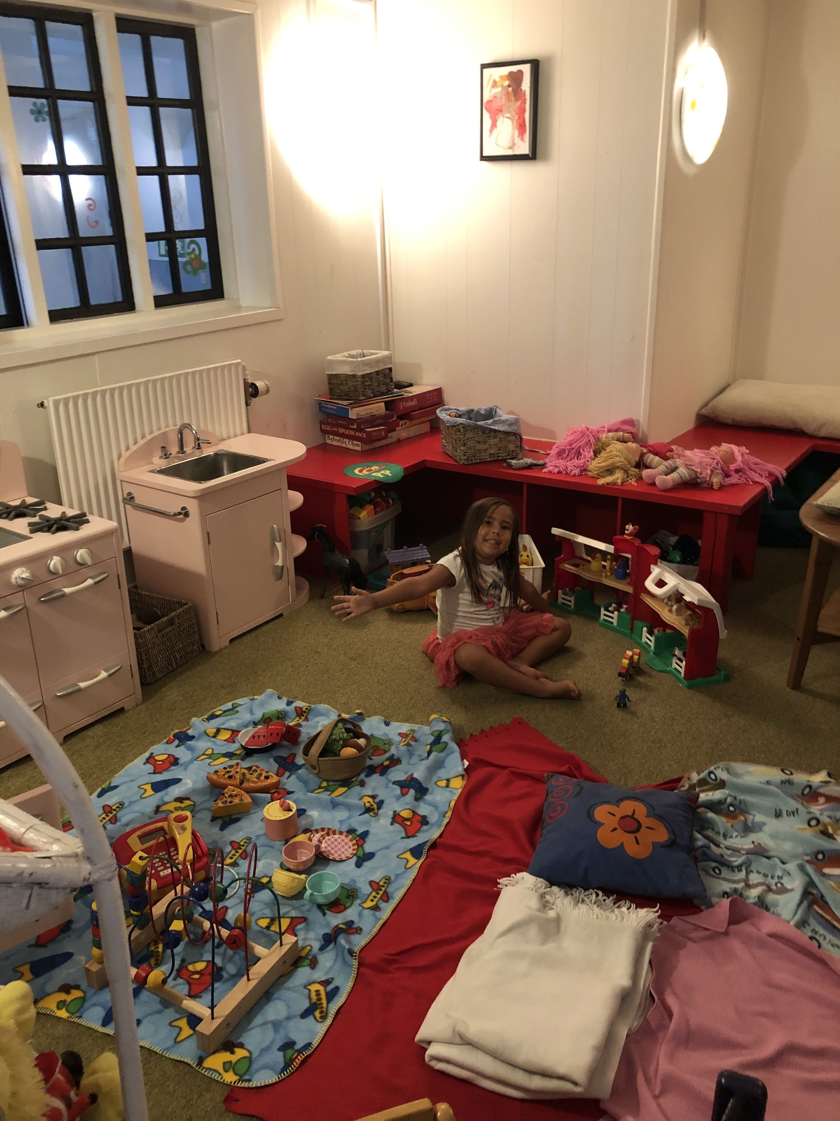 Un cuarto de juegos infantil con una mesa roja para jugar, una pequeña cocina y una ventana con persianas.