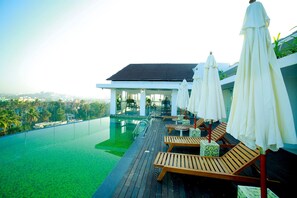 An infinity pool with wooden lounge chairs and umbrellas, a covered seating area, and a view of the cityscape. - Vivin Luxury Suites (Thiruvananthapuram)