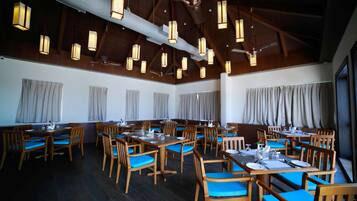 A spacious dining area with multiple tables and chairs, wooden beams on the ceiling, and hanging pendant lights.