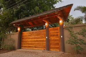 A wooden gate with a metal roof structure, surrounded by a gravel driveway and a wall with vegetation. - Hermandad, Short Walk to Surfing Beach | Family Retreat, Gorgeous 4 BDR home located very close to the Beach Dog Cafe  (Nosara)