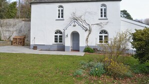 Una casa blanca de dos pisos amb un teulat gris, un petit porxo davanter i un jardí amb diverses plantes i una estructura de fusta.
