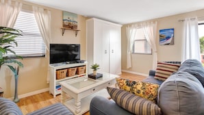 A living room with a television, sofa, and a coffee table. There are curtains, a plant, and a painting on the wall. - 🌴 Gulf Winds Studio — Balcony Views & Sleep Number Comfort by the Beach (St. Pete Beach)