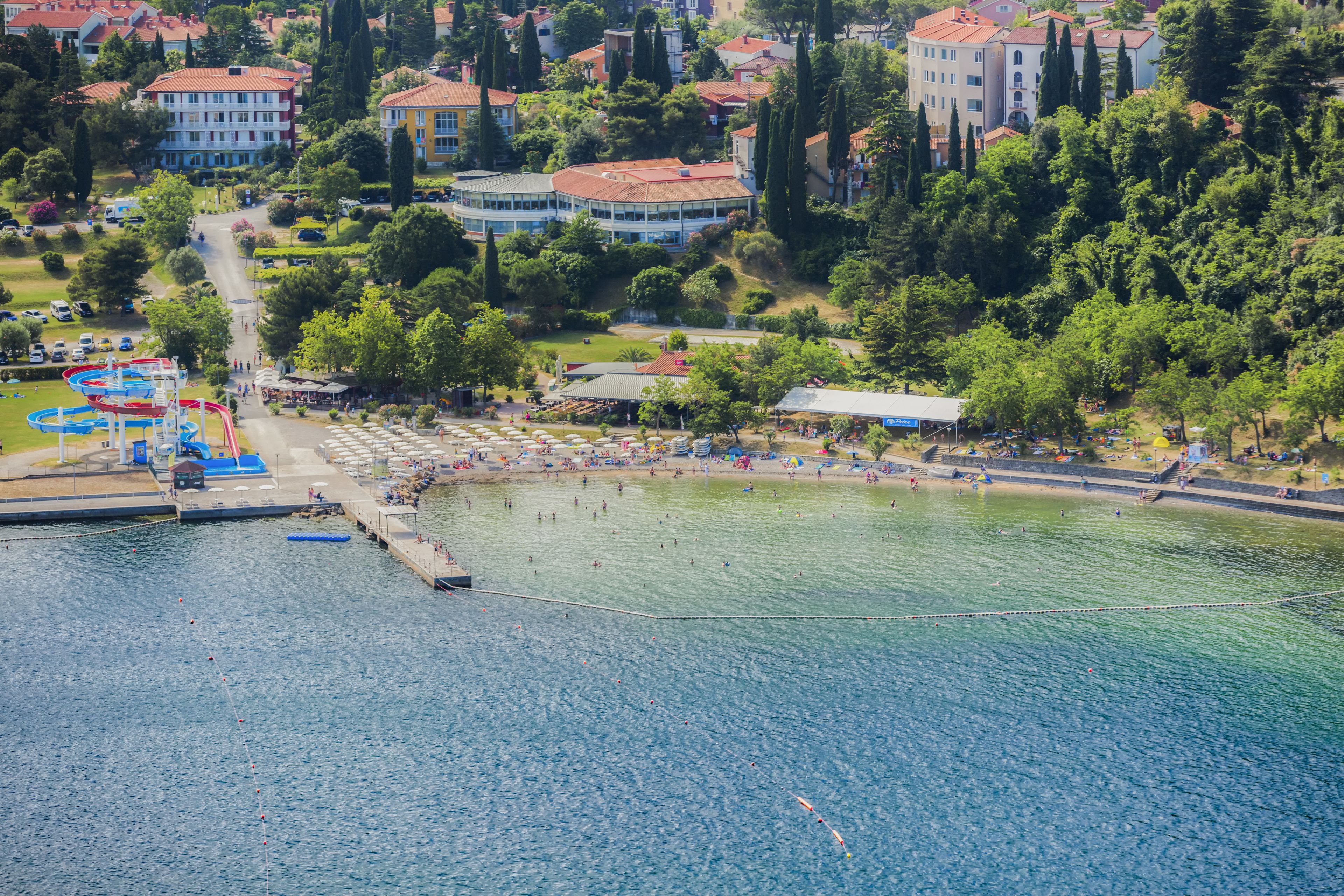 An aerial view of a coastal area with a beach, water sports facilities, and surrounding buildings.
