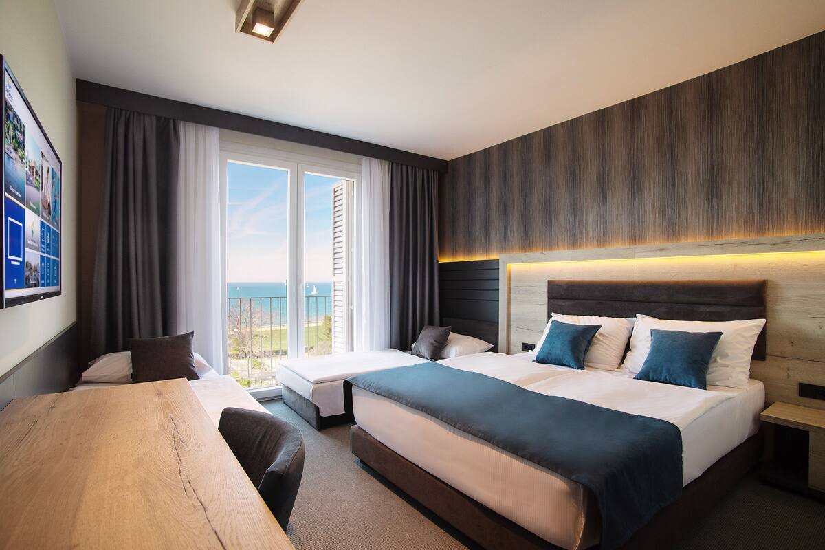 Quarto de hotel com duas camas, uma escrivaninha de madeira e uma janela grande com vista para o mar.