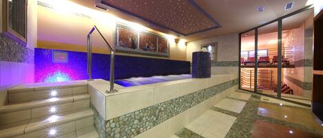 Un espace spa doté d’un sol carrelé en pierre, d’un élément décoratif aquatique illuminé de lumière bleue et d’un coin salon offrant une vue sur l’espace spa.