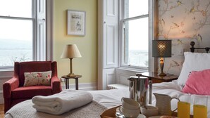 Ein Hotelzimmer mit Bett, Sessel, Tablett mit Frühstücksgebäck und Meerblick durch große Fenster.
