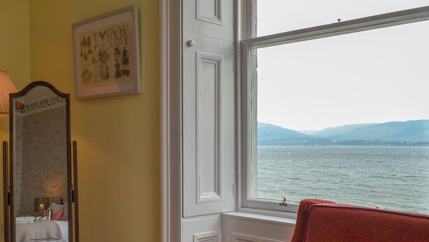 Ein Zimmer mit großem Fenster mit Blick auf ein Gewässer, einem Spiegel und einem Sessel mit Blumenkissen.