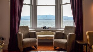 Ein Zimmer mit Erkerfenster, das auf ein Gewässer und entfernte Landschaft blickt. Zwei Sessel und ein kleiner Tisch mit Fernglas stehen darin.
