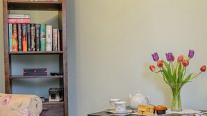 A room with a bookshelf, a floral armchair, a table with a teapot, cups, and a plate of pastries. - 1 bedroom accommodation in Cawston, near Aylsham (Cawston, near Aylsham)