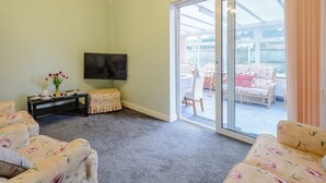 A living room with a flat-screen TV, floral-patterned sofas, a small table with a teapot and tulips, and a sliding glass door leading to an outdoor patio with a chair and a table. - 1 bedroom accommodation in Cawston, near Aylsham (Cawston, near Aylsham)