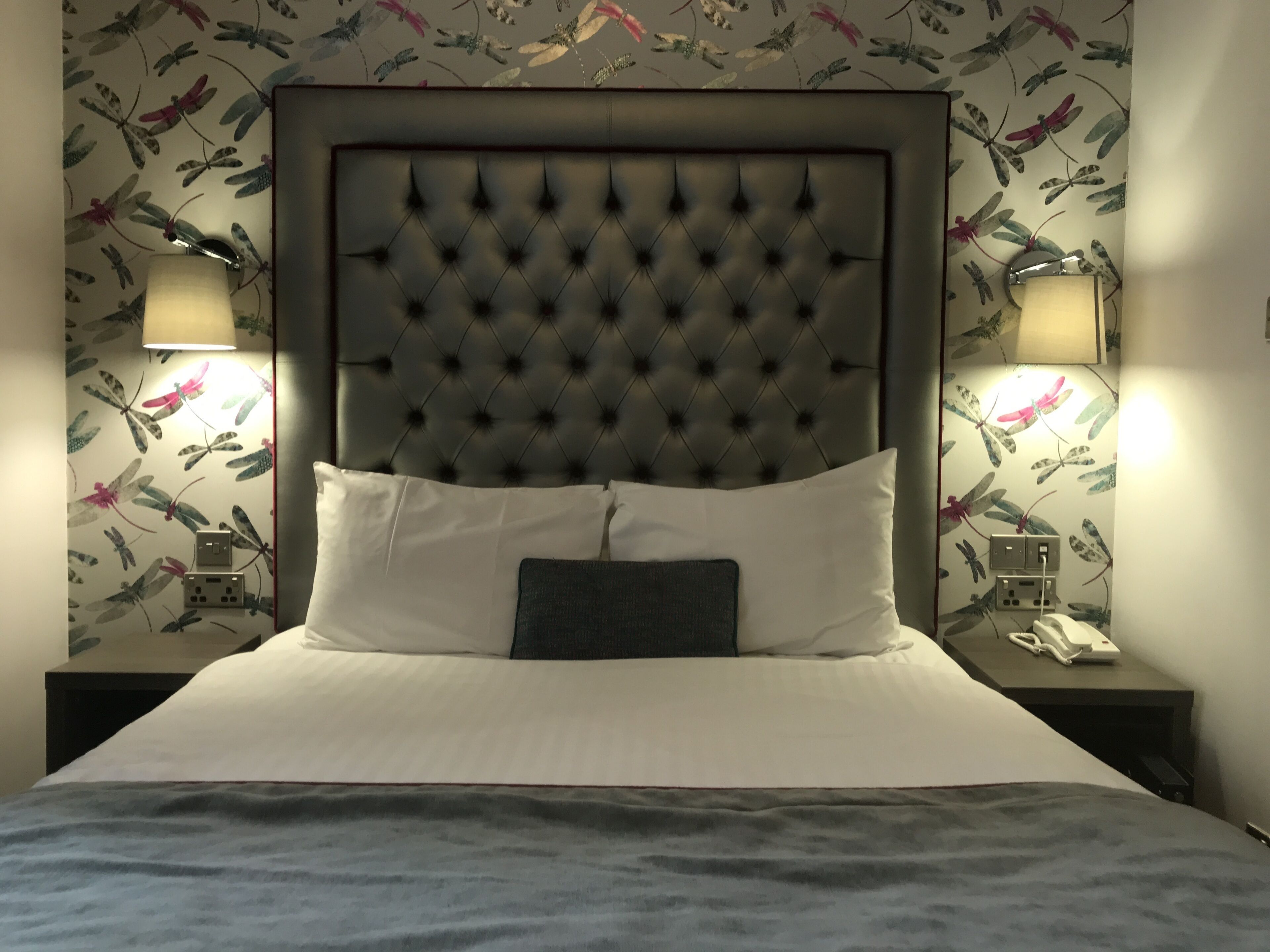 A hotel room with a tufted headboard, white bedding, and floral wallpaper.